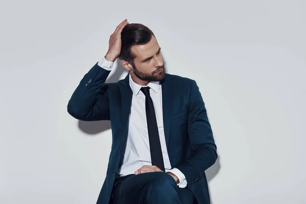 Momento para pensar. Hombre joven guapo ajustando su peinado mientras está sentado sobre fondo gris —  Fotos de Stock