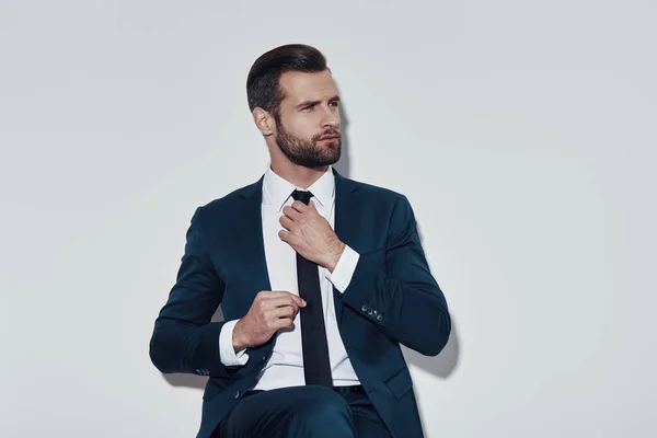 Prestando atención a cada detalle. Guapo joven ajustando su corbata mientras está sentado sobre fondo gris — Foto de Stock