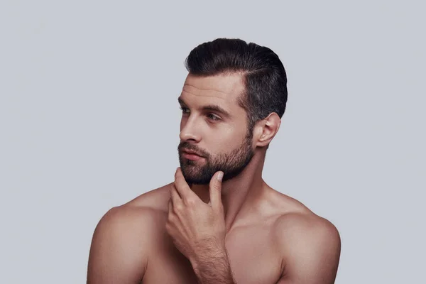 Hora de afeitarse. Joven guapo tocándose la barba mientras está de pie sobre un fondo gris —  Fotos de Stock