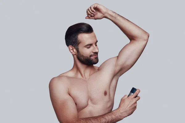 Freshness. Handsome young man applying deodorant and smiling while standing against grey background — Stock Photo, Image