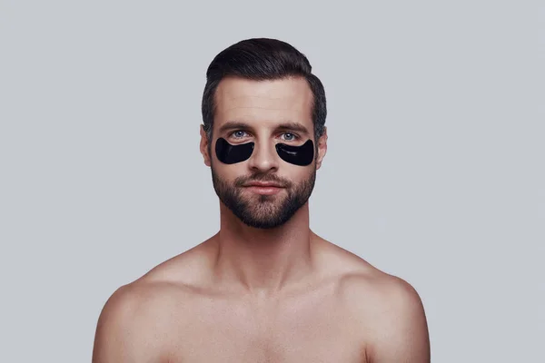 Tratamiento de belleza. Hombre joven guapo que aplica parche de ojo médico y sonríe mientras está de pie sobre fondo gris —  Fotos de Stock