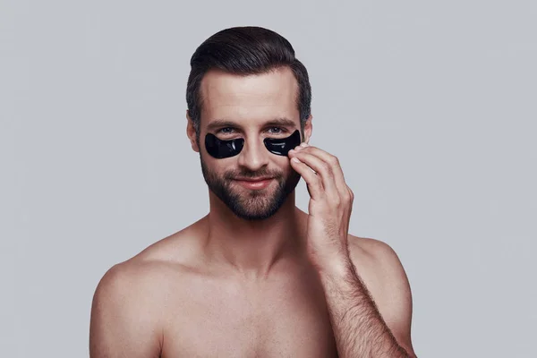 Cuidando su piel. Hombre joven guapo que aplica parche de ojo médico y sonríe mientras está de pie sobre fondo gris —  Fotos de Stock