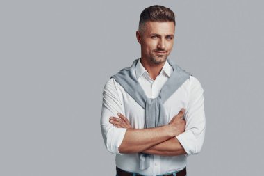 Tarzına güveniyor. Yakışıklı genç adam kameraya bakıyor ve gri arka planda dururken gülümsüyor