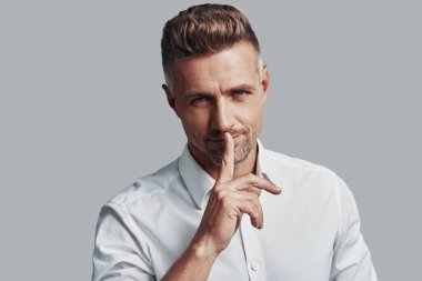 Sırrımı sakla! Yakışıklı genç adam kamera bakıyor ve gri arka planda ayakta iken dudaklarında parmak tutarak