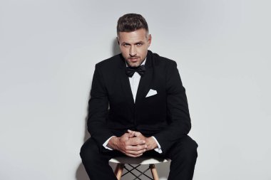 Kendinden emin bir görünüm. Gri arka plan aleyhine otururken kameraya bakarak tam takım elbise ve papyon yakışıklı genç adam