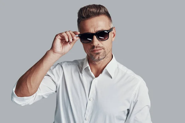 In his own style. Handsome young man looking at camera and adjusting sunglasses while standing against grey background — Stock Photo, Image