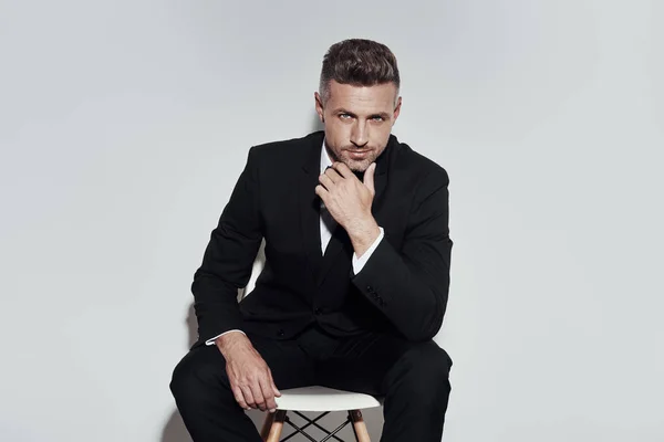 Confident and charming. Handsome young man in full suit and bow tie looking at camera while sitting against grey background — Stock Photo, Image