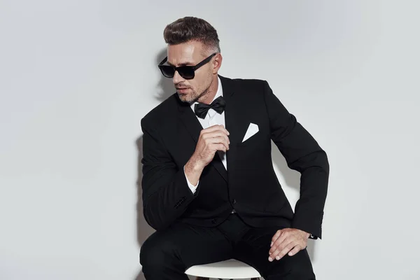 Luxury look. Handsome young man in full suit and bow tie looking away while sitting against grey background — Stock Photo, Image