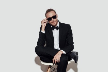 Mükemmel bir adam. Gri arka plan aleyhine otururken kameraya bakan ve güneş gözlüğü ayarlayan tam takım elbiseli Charming genç adam