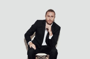 Genç ve kendinden emin. Takım elbiseli yakışıklı genç adam gri arka planda otururken kameraya bakıyor.