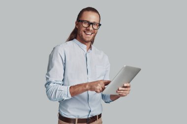 Modern teknolojiler. Yakışıklı genç adam dijital tablet kullanarak çalışan ve gri arka plan karşı dururken gülümseyerek