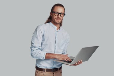Çözüm aranıyor. Yakışıklı genç adam dizüstü bilgisayar kullanarak çalışan ve gri arka plan karşı dururken gülümseyerek