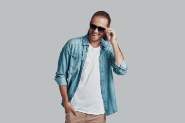 Tek kelimeyle büyüleyici. Yakışıklı genç adam güneş gözlüğü ayarlama ve gri arka plan karşı dururken gülümseyerek