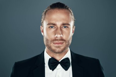 Kendinden emin bir görünüm. Gri arka plan aleyhine dururken kameraya bakan ve gülümseyen papyonlu büyüleyici genç adam