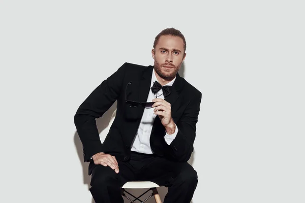 Heartbreaker. Charming young man in full suit looking at camera while sitting against grey background — Stock Photo, Image