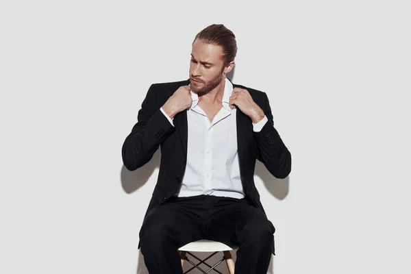 Estilo perfecto. Hombre joven guapo en traje completo mirando hacia otro lado y ajustando la chaqueta mientras está sentado sobre un fondo gris — Foto de Stock