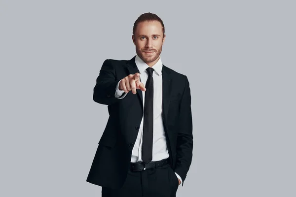I need you! Handsome young man in full suit looking at camera and pointing you while standing against grey background