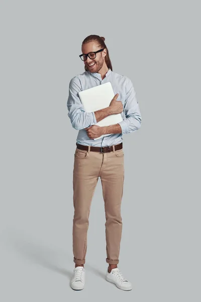 Νέος και έξυπνος. Πλήρες μήκος του όμορφος νεαρός άνδρας μεταφέρουν φορητό υπολογιστή και χαμογελώντας, ενώ στέκεται πάνω από γκρι φόντο — Φωτογραφία Αρχείου