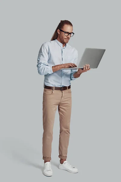 Esperto in affari. Piena lunghezza di bel giovane uomo che lavora con il computer portatile mentre in piedi su sfondo grigio — Foto Stock