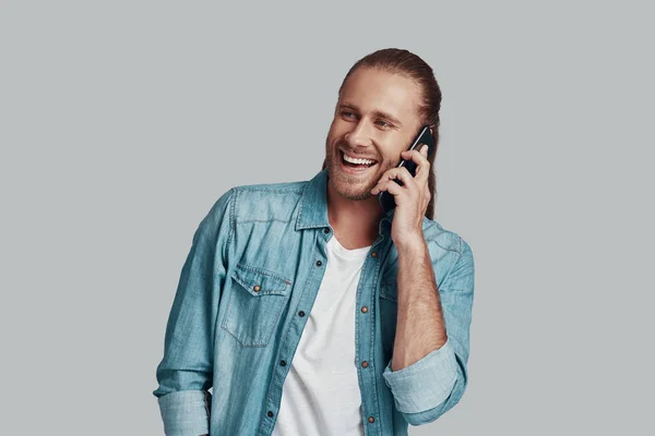 Fico feliz em ouvir-te! Bonito jovem falando no telefone inteligente e sorrindo enquanto está de pé contra fundo cinza — Fotografia de Stock