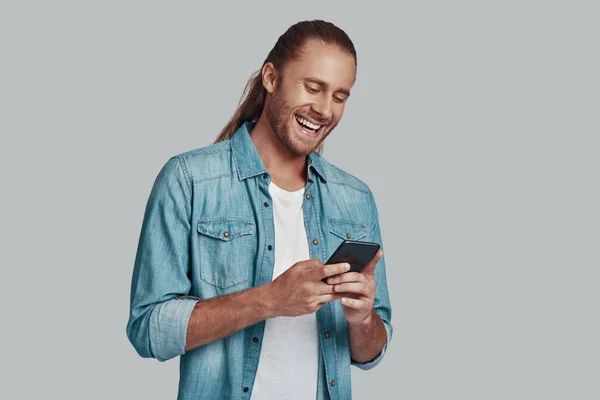 Social gathering. Handsome young man using smart phone and smiling while standing against grey background