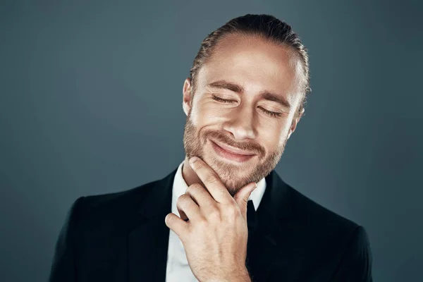 Affascinante giovane uomo in papillon sorridente e tenere la mano sul mento mentre in piedi sullo sfondo grigio — Foto Stock