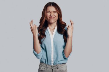 Bir mucize bekliyorum. Gri arka planda dururken parmaklarını çapraz ve gözleri kapalı tutan akıllı rahat giyim genç kadın