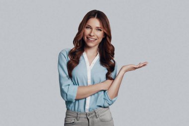 Şuna bak! Çekici genç kadın gri arka plan karşı dururken kamera bakıyor ve kopyalama alanı işaret