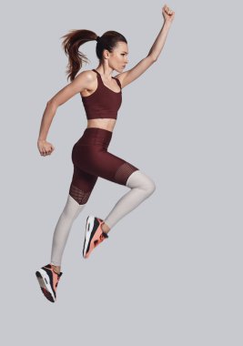 Hareket etmeye devam et! Gri arka plan aleyhine egzersiz yaparken spor giyim atlama çekici genç kadın tam uzunlukta