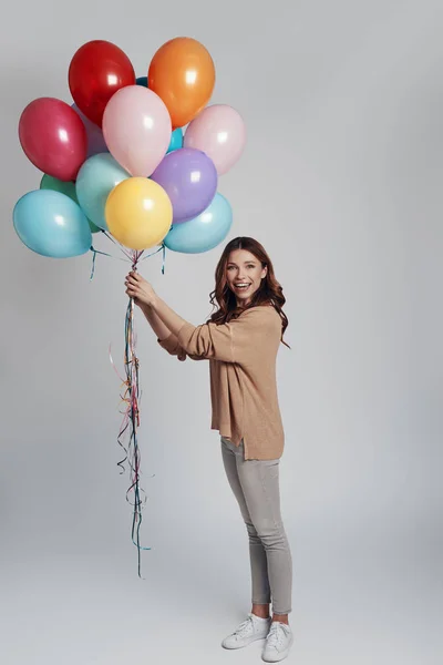 Accidenti! Piena lunghezza di attraente giovane donna che tiene palloncini e sorride mentre in piedi contro lo sfondo grigio — Foto Stock