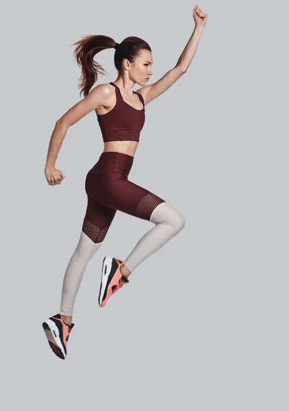 Продовжуйте рухатися! Повна довжина привабливої молодої жінки в спортивному одязі стрибає під час тренувань на сірому фоні — стокове фото
