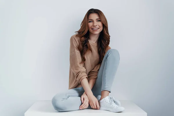 Pura perfección. Atractiva joven mujer mirando a la cámara y sonriendo mientras se sienta sobre un fondo gris — Foto de Stock