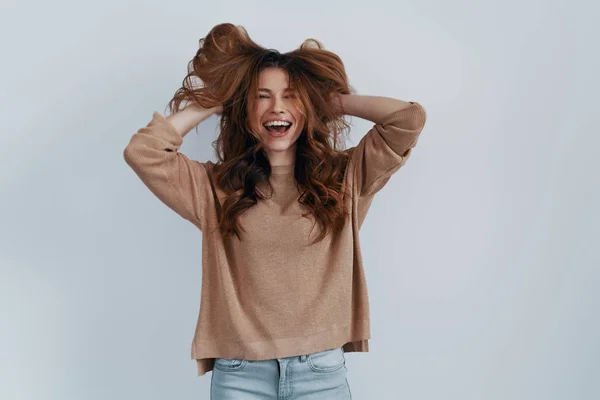 Penteado perfeito. Mulher bonita mantendo as mãos no cabelo e sorrindo enquanto está de pé contra fundo cinza — Fotografia de Stock