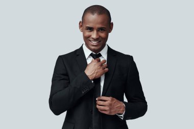 Zarif adam. Kravatı ayarlayan ve gri arka planda dururken gülümseyerek kameraya bakan formalgiyimli yakışıklı genç Afrikalı adam