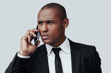 Tam konsantrasyon. Düşünceli genç Afrikalı adam formalofa akıllı telefonda konuşurken ve gri arka plan aleyhine dururken kaşlarını çatma