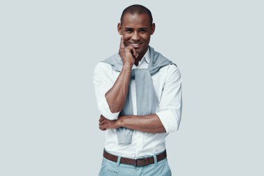 Çok çekici. Yakışıklı genç Afrikalı adam kameraya bakıyor ve gri arka plan aleyhine dururken gülümsüyor
