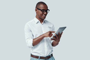  Yeni tableti inceliyorum. Yakışıklı genç Afrikalı adam dijital tablet kullanarak ve gri arka plan karşı dururken gülümseyerek 
