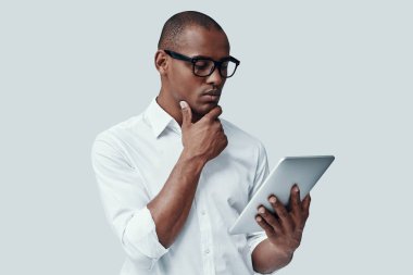 Ya... Yakışıklı genç Afrikalı adam dijital tablet kullanarak ve gri arka plan karşı dururken çene üzerinde el tutmak