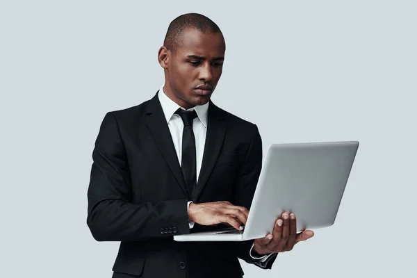 Pronto para fazer negócios. Jovem africano em trajes formais que trabalha usando o computador enquanto está de pé contra fundo cinza — Fotografia de Stock