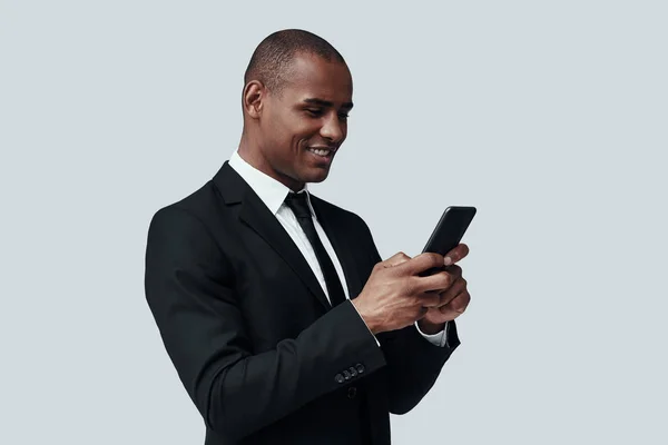 Sempre disponível. Encantador jovem africano em formalwear usando telefone inteligente e sorrindo enquanto está de pé contra fundo cinza — Fotografia de Stock