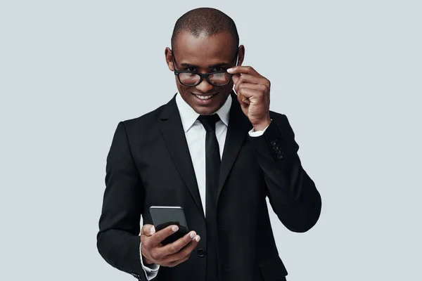 Ειδικός στις επιχειρήσεις με αυτοπεποίθηση. Γοητευτικός νέος Αφρικανός άντρας σε τυποφόρα χρησιμοποιώντας έξυπνο τηλέφωνο και χαμογελώντας ενώ στέκεται ενάντια σε γκρίζο φόντο — Φωτογραφία Αρχείου