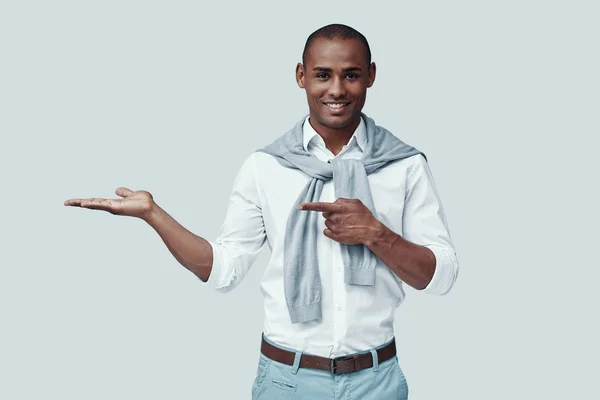 Look here! Handsome young African man looking at camera and smiling while standing against grey background