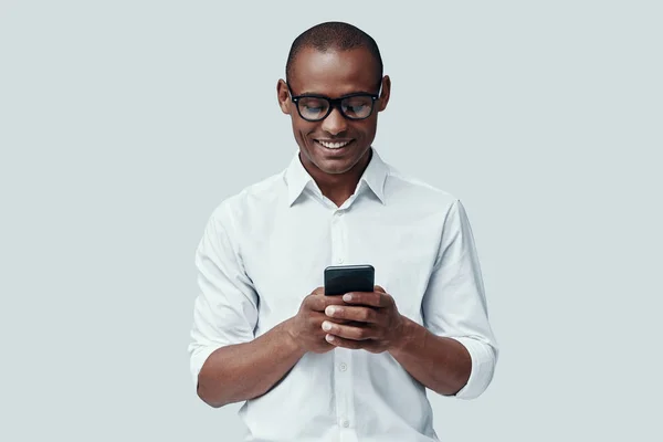 Mensajería de texto. Hombre africano joven guapo usando el teléfono inteligente mientras está de pie sobre fondo gris —  Fotos de Stock