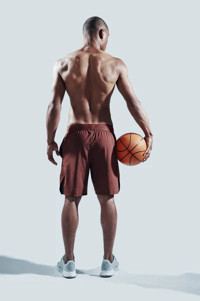 In a great shape. Full length rear view of handsome young African man in sports clothing carrying basketball ball while standing against grey background