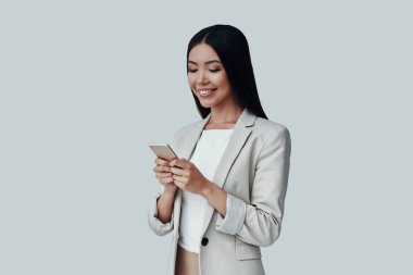 Her zaman kullanılabilir. Çekici genç Asyalı kadın akıllı telefon kullanarak ve gri arka plan karşı dururken gülümseyerek