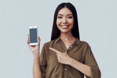 Bir bak! Çekici genç Asyalı kadın akıllı telefon işaret ve gri arka plan karşı dururken gülümseyerek