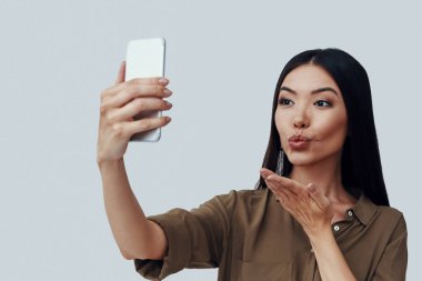 Bir öpücük üfleme. Çekici genç Asyalı kadın selfie çekerken ve gri arka planda dururken bir öpücük üfleme