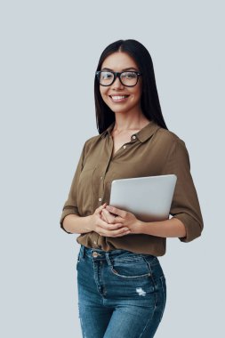 Kendine güvenen ve akıllı. Çekici genç Asyalı kadın kameraya bakıyor ve gri arka planda dururken gülümsüyor