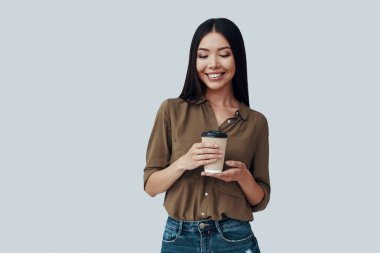 Kahvenin tadını çıkarıyorum. Gri arka planda dururken gülümseyen güzel genç Asyalı kadın