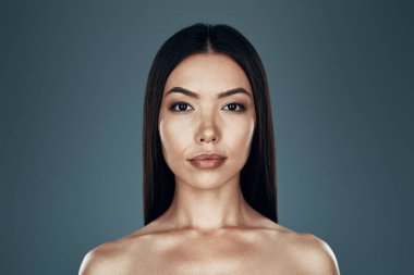 Doğal güzelliklerle parlıyor. Gri arka planda dururken kameraya bakan güzel genç Asyalı kadın